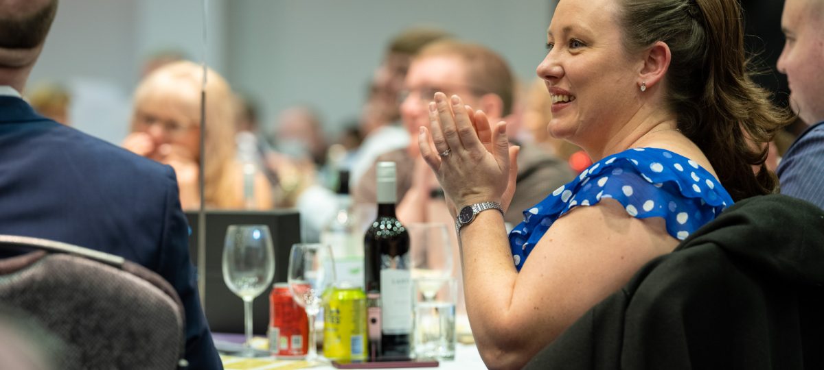 Image shows a lady in a blue polka dot dress sat at a round table with friends. They are clapping.